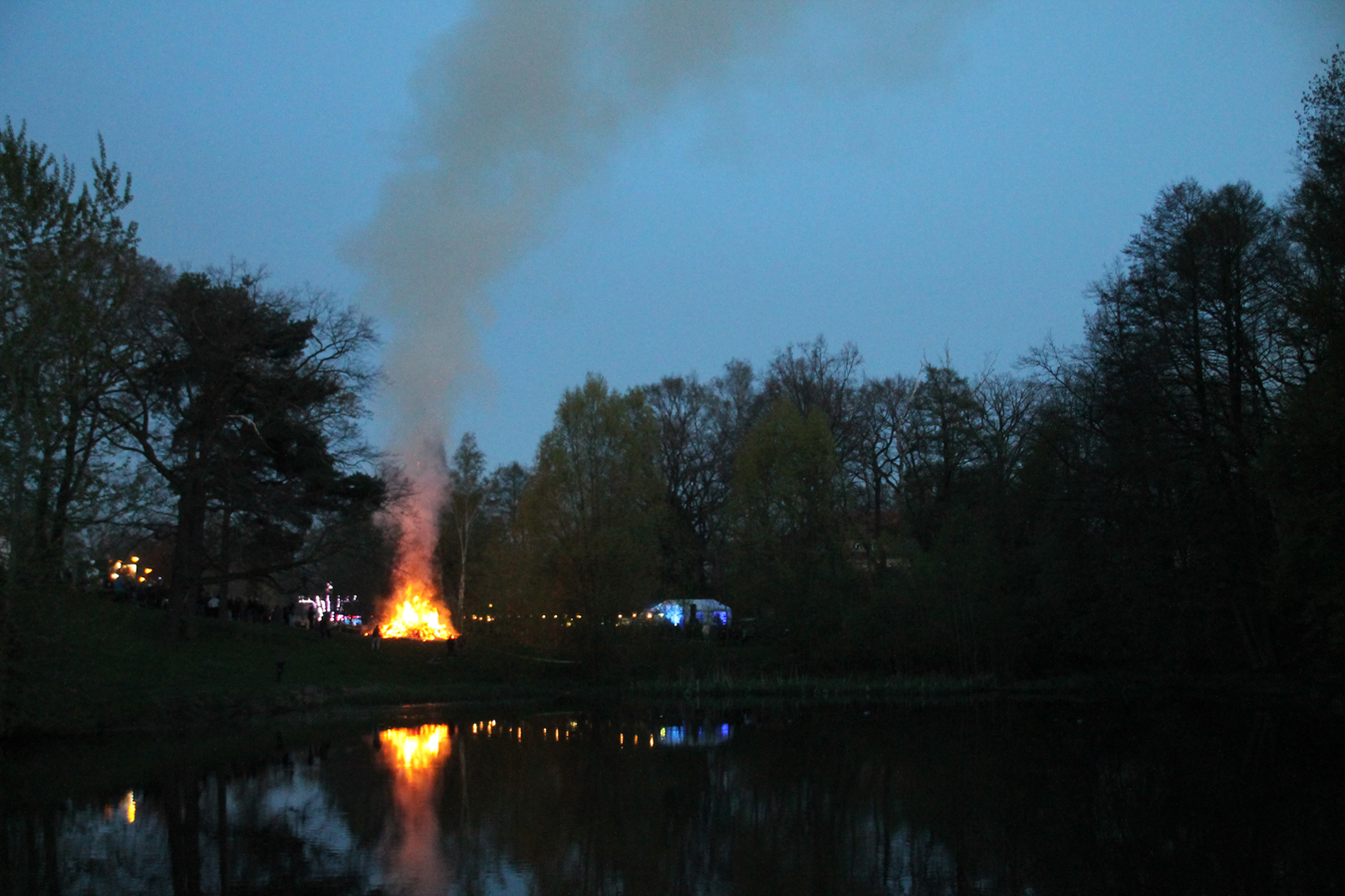 Großes Lagerfeuer lodert am Ufer eines Sees bei Dämmerung.